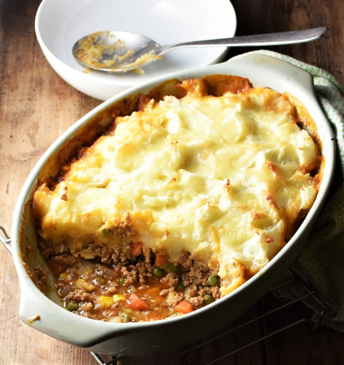 Side view of turkey cottage pie in oval green dish with white bowl and spoon in background.