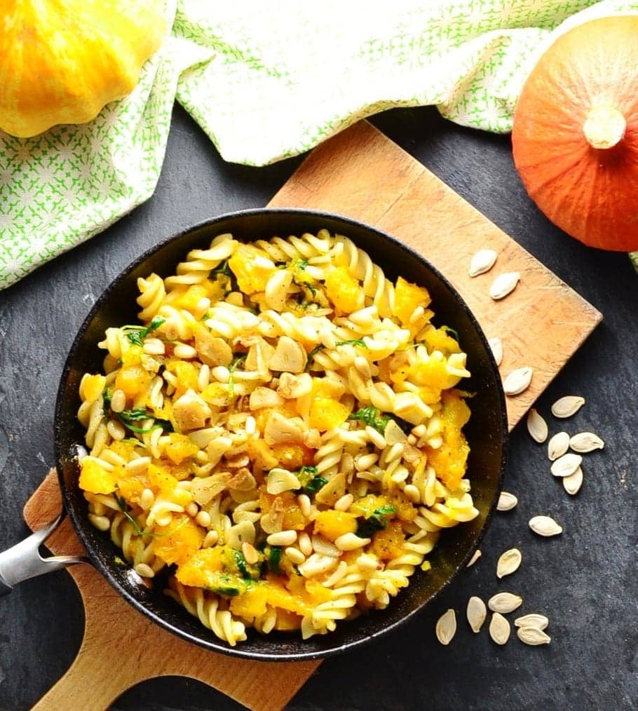 Top down view of fussili pasta with pumpkin pieces, garlic slices and pine nuts inside skillet on top of wooden board, with green cloth, pumpkin seeds and squash in background.