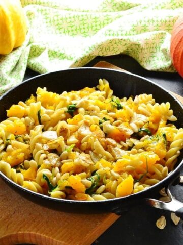 Side view of chopped pumpkin with fussili pasta, arugula, garlic slices and nuts on top of wooden board, with green cloth and pumpkin in background.