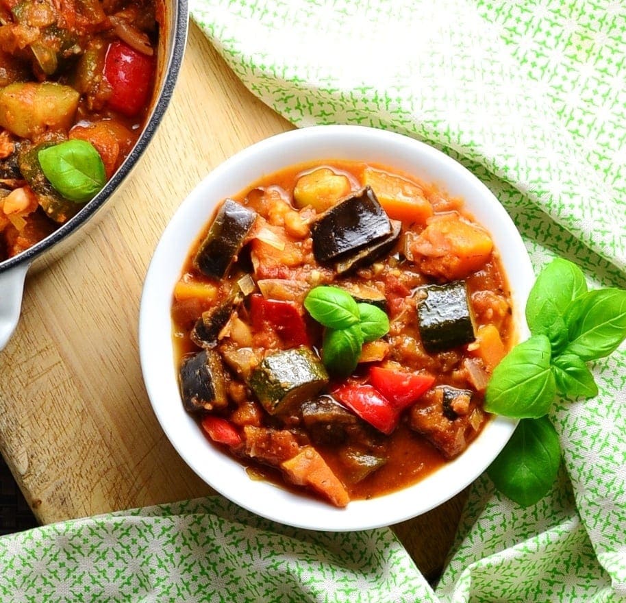 Top down view of vegetable stew in white bowl with green cloth around it on wooden board with stew in pot in top left corner.