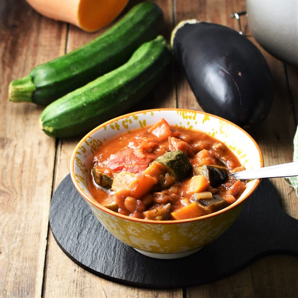 Chunky vegetable casserole in yellow patterned bowl with spoon, eggplant, 2 zucchinis in background.