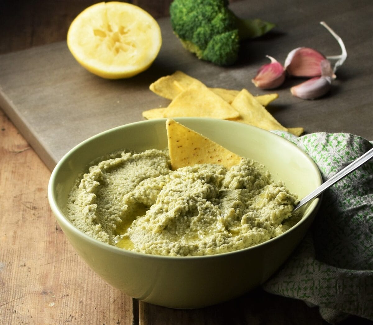 Broccoli dip in green bowl with spoon, nachos, lemon, broccoli and garlic in background.