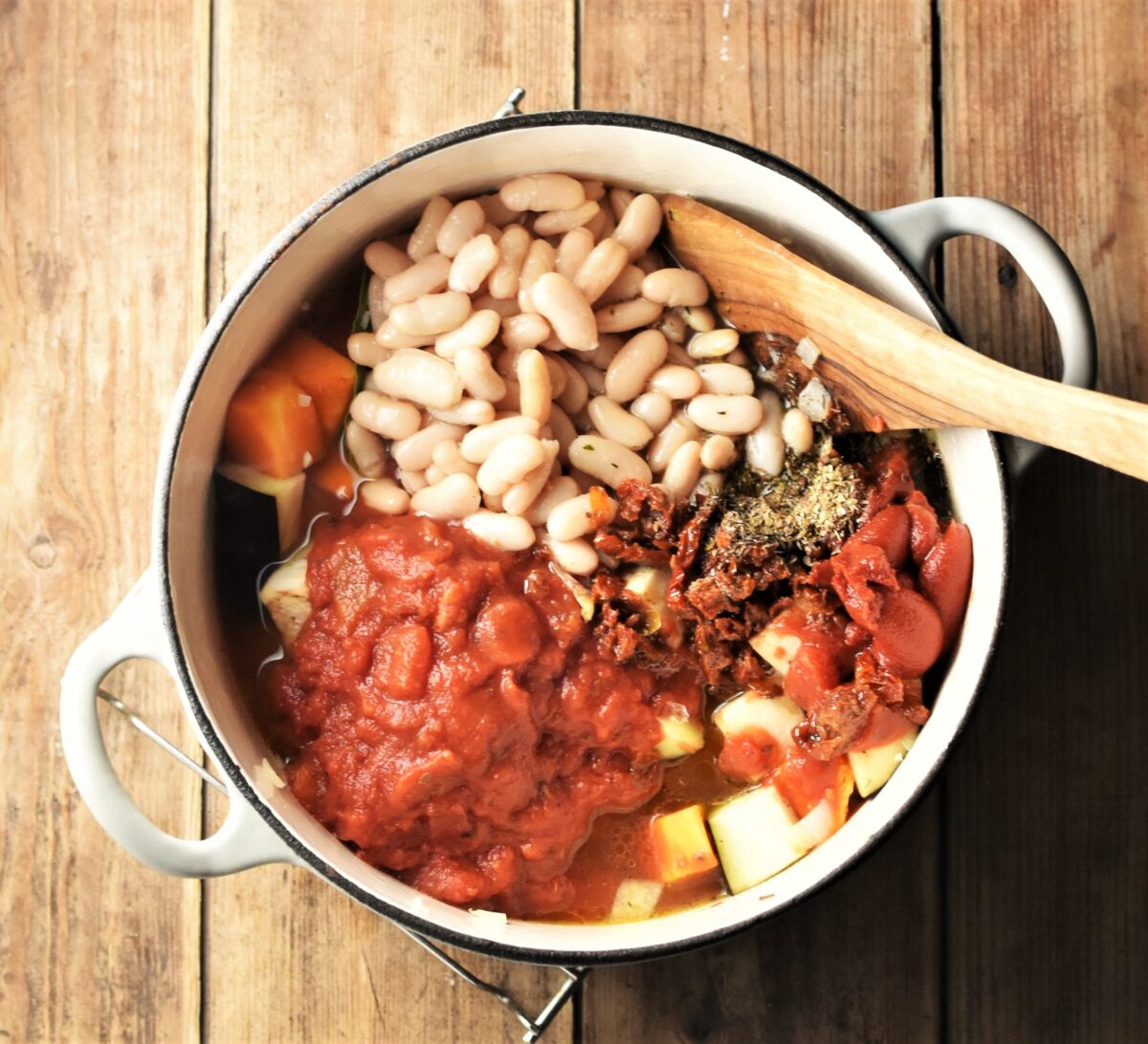 Chopped tomatoes, white beans, tomato paste and wooden spatula in large white pot.