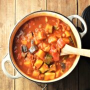 Chunky vegetable stew in tomato sauce in large white post with wooden spoon.