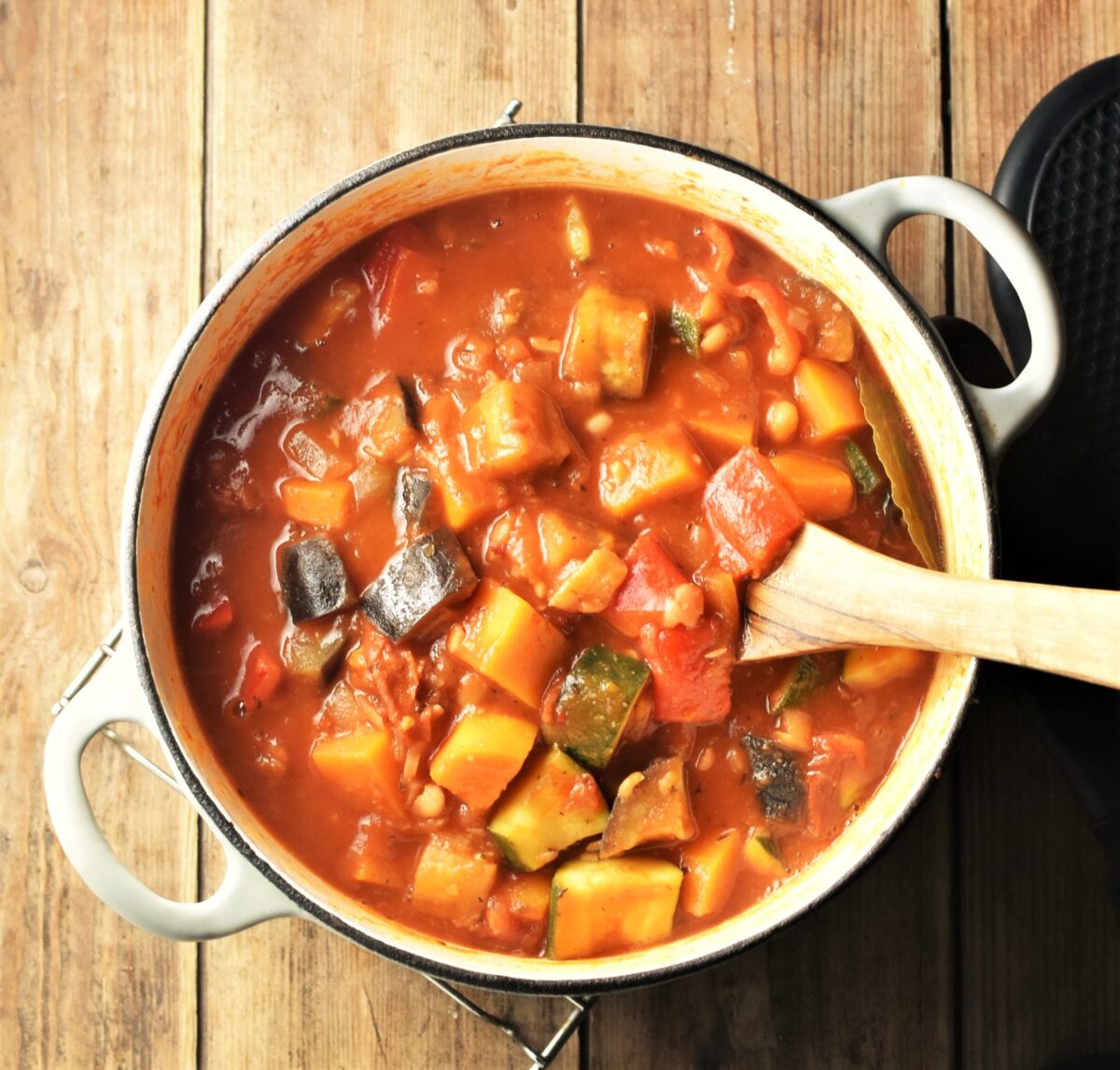 Chunky vegetable stew in tomato sauce in large white post with wooden spoon.