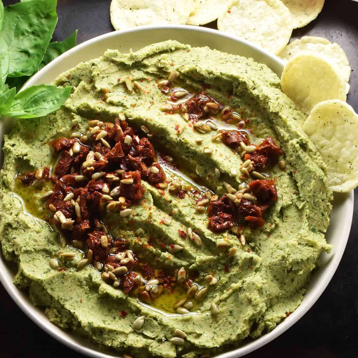 Close-up view of creamy broccoli bean dip topped with sundried tomatoes with crisps in background.