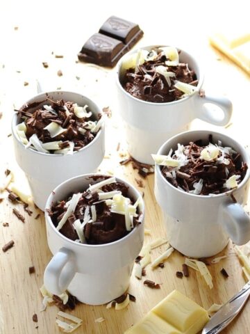 Cheesecake pots with white and dark chocolate shavings, chocolate pieces and spoon on wooden surface.