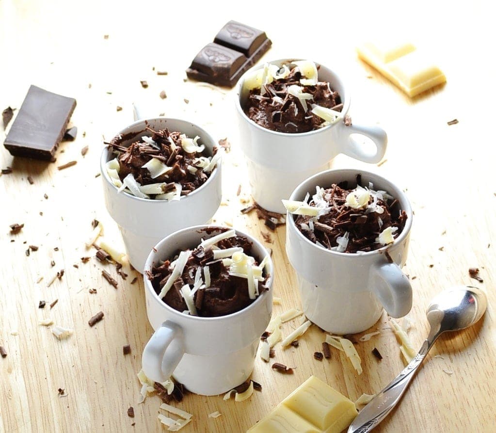 Chocolate cheesecake pots with chocolate shavings, chocolate pieces and spoon on wooden surface.
