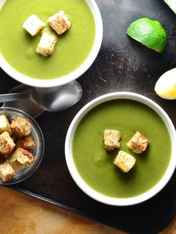 Top down view of split pea soup in 2 white bowls with croutons, lime wedges, spoons and dish of croutons on oven tray.