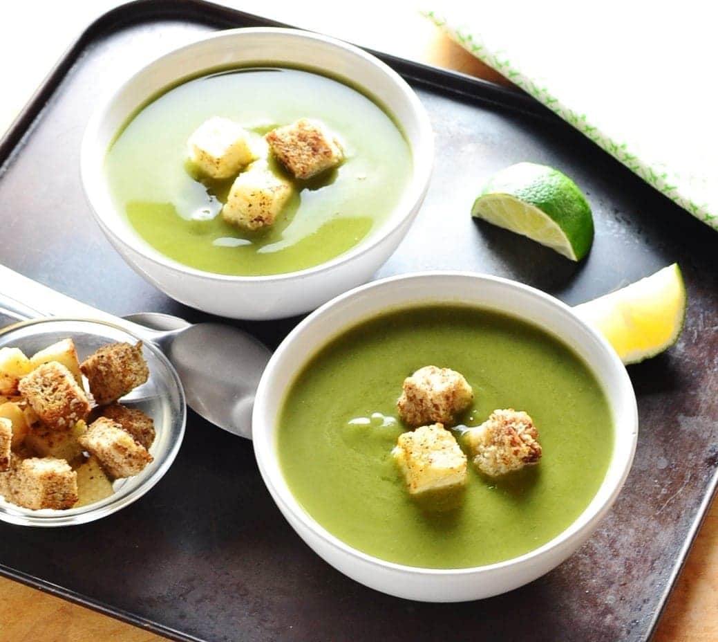 Green pea soup in 2 bowls with croutons, lime wedges, croutons in small dish and spoons on dark oven tray.