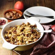 Vegetarian stuffing in white oval dish with spoon and red-and-white cloth to the right,plates and forks, chestnuts and apple in background.