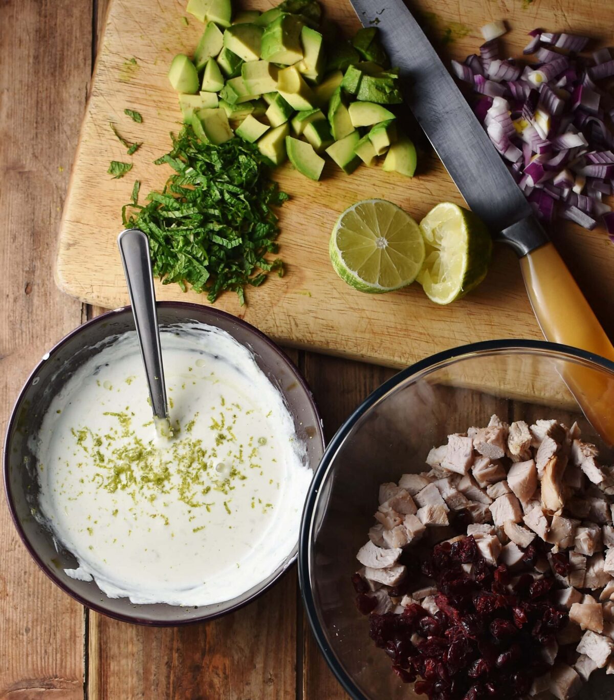 Yogurt in bowl with spoon, chopped turkey and dried cranberries in another bowl, lime, chopped herbs, avocado, red onions and knife in background.