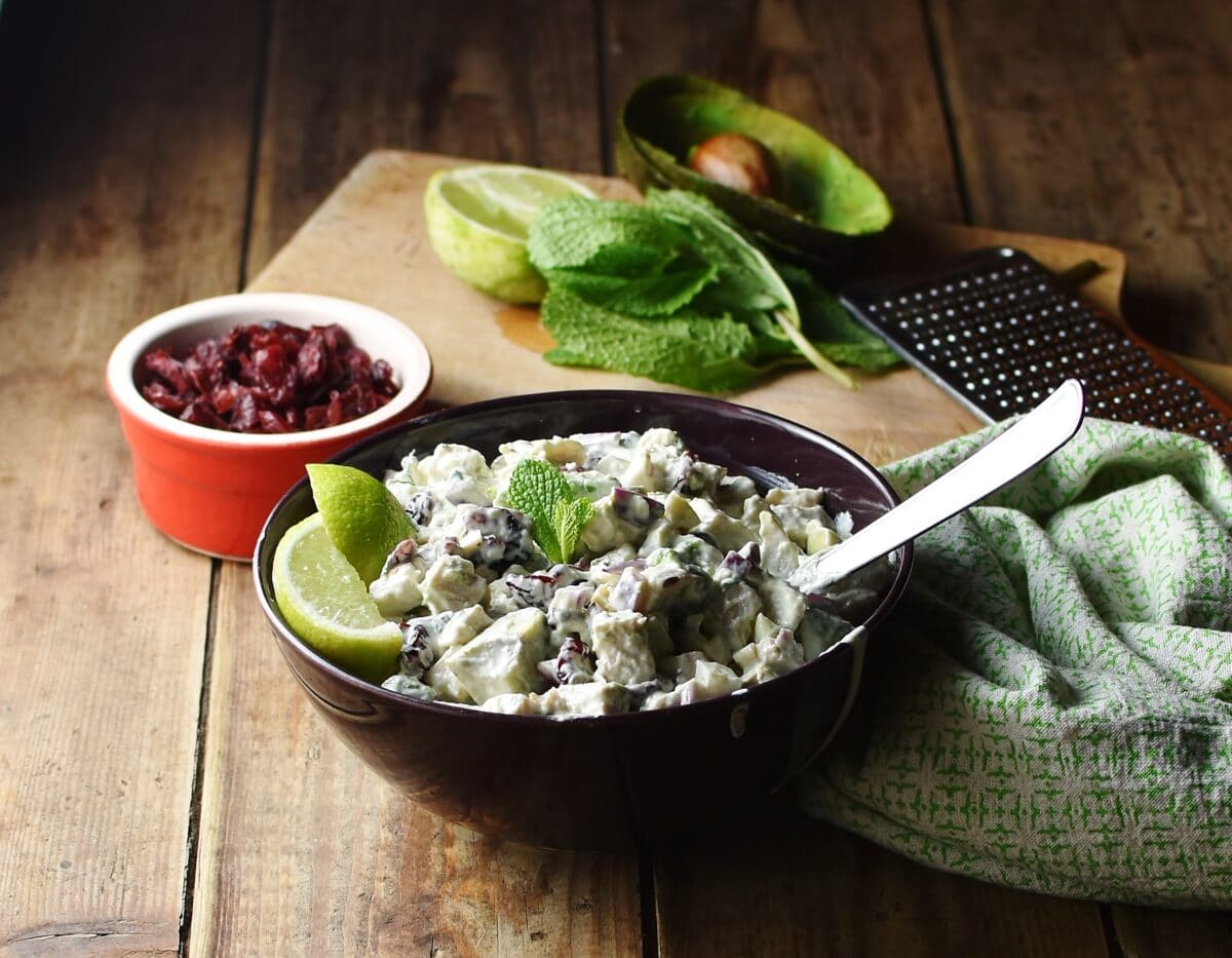 Side view of creamy turkey salad with lime wedges and spoon in purple bowl, with dried cranberries in red dish, mint leaves, lime, avocado peel and green cloth in background.