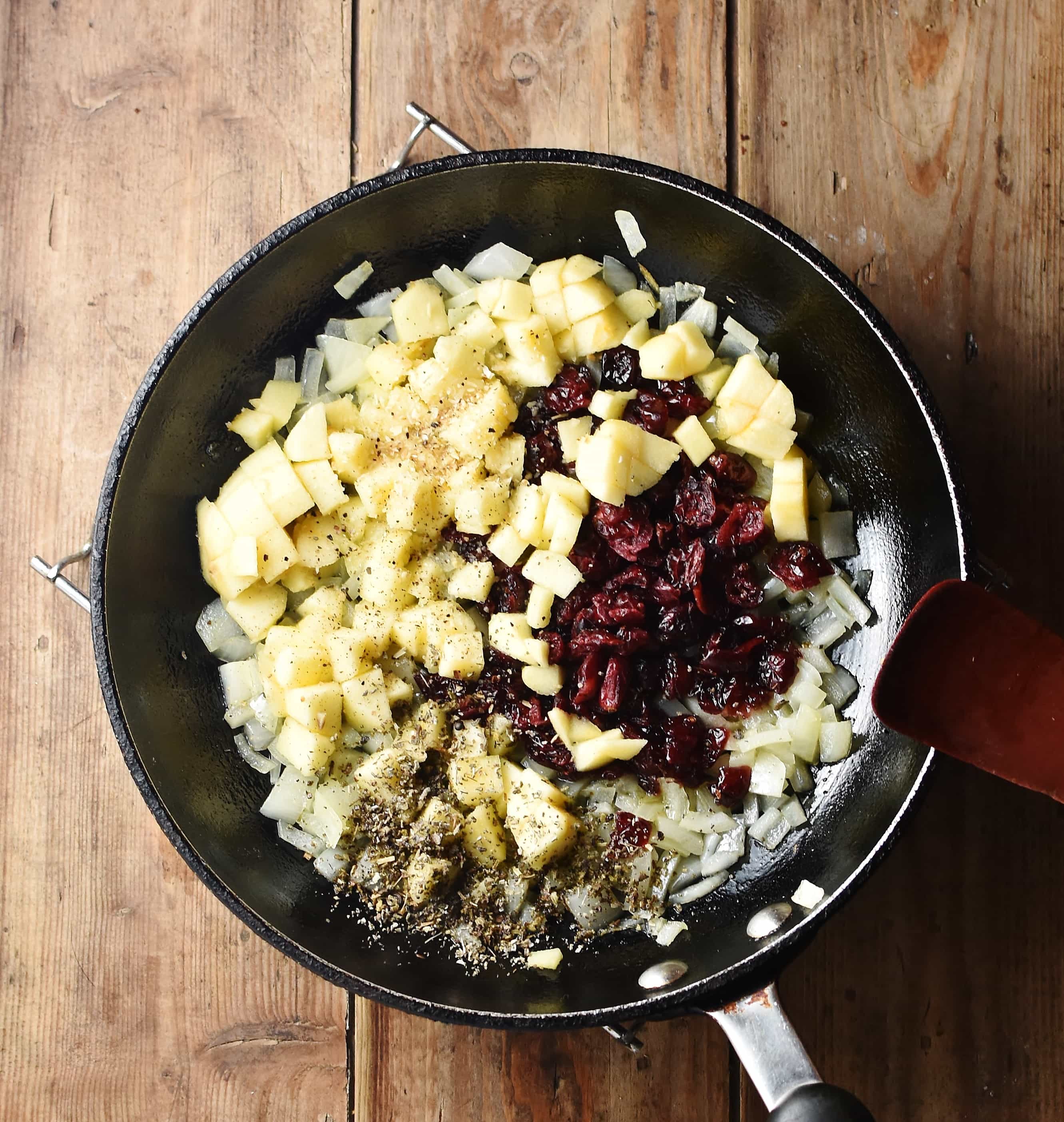 Chopped apple, cranberries, onions and spices in skillet with red spatula ti the right.