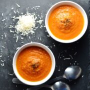 Top down view of creamy tomato soup in 2 white bowls with grated cheese and 2 spoons in background.