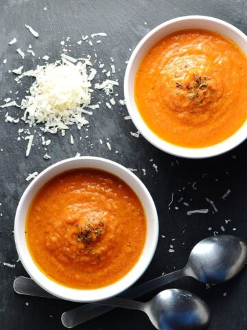 Top down view of creamy tomato soup in 2 white bowls with grated cheese and 2 spoons in background.