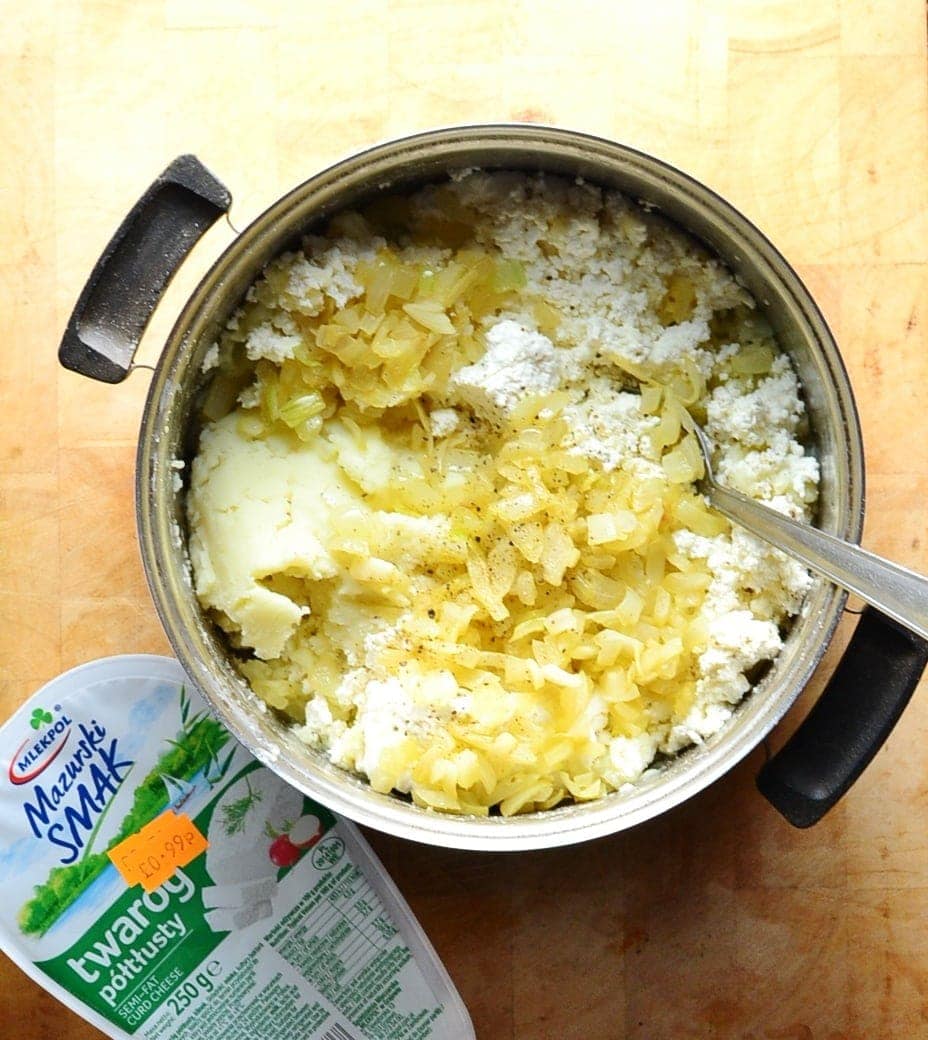 Top down view of Polish cheese and potato pierogi filling in saucepan with spoon and Polish cheese on top of wooden surface.