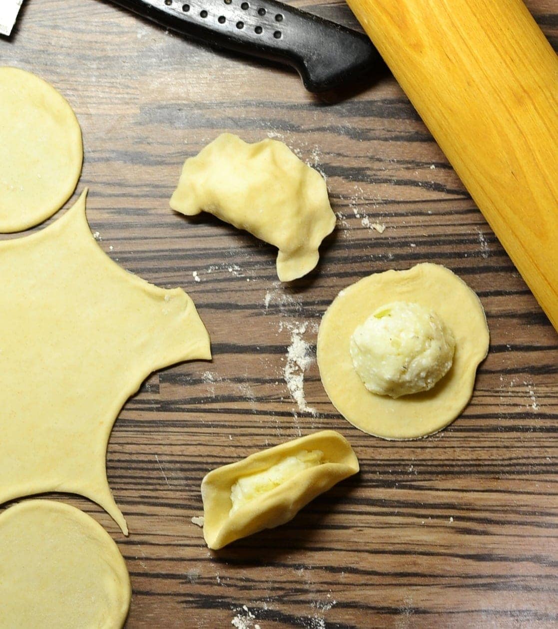 Rolled out dough and partly made pierogi with rolling pin in top right corner on top of wooden surface.