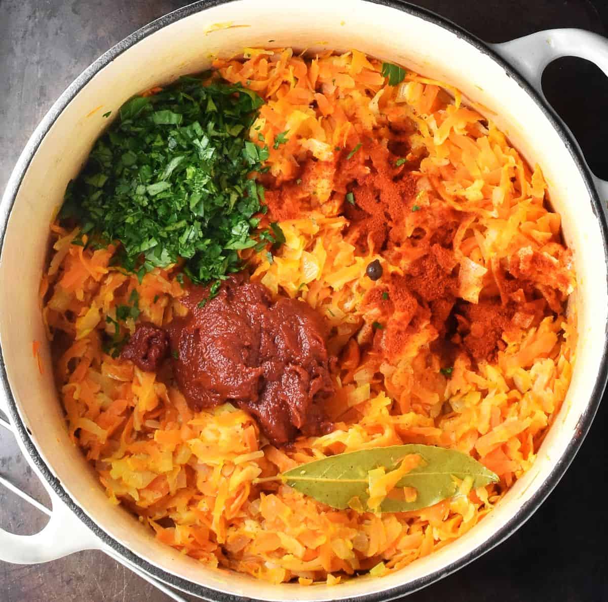 Top down view of thick vegetable stew with tomato paste, chopped parsley, paprika and bay on top in white pot.