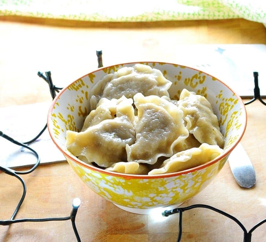 Sauerkraut pierogi in yellow-and-white patterned bowl with Christmas themed coasters, Christmas lights and green cloth in background on light wooden surface.