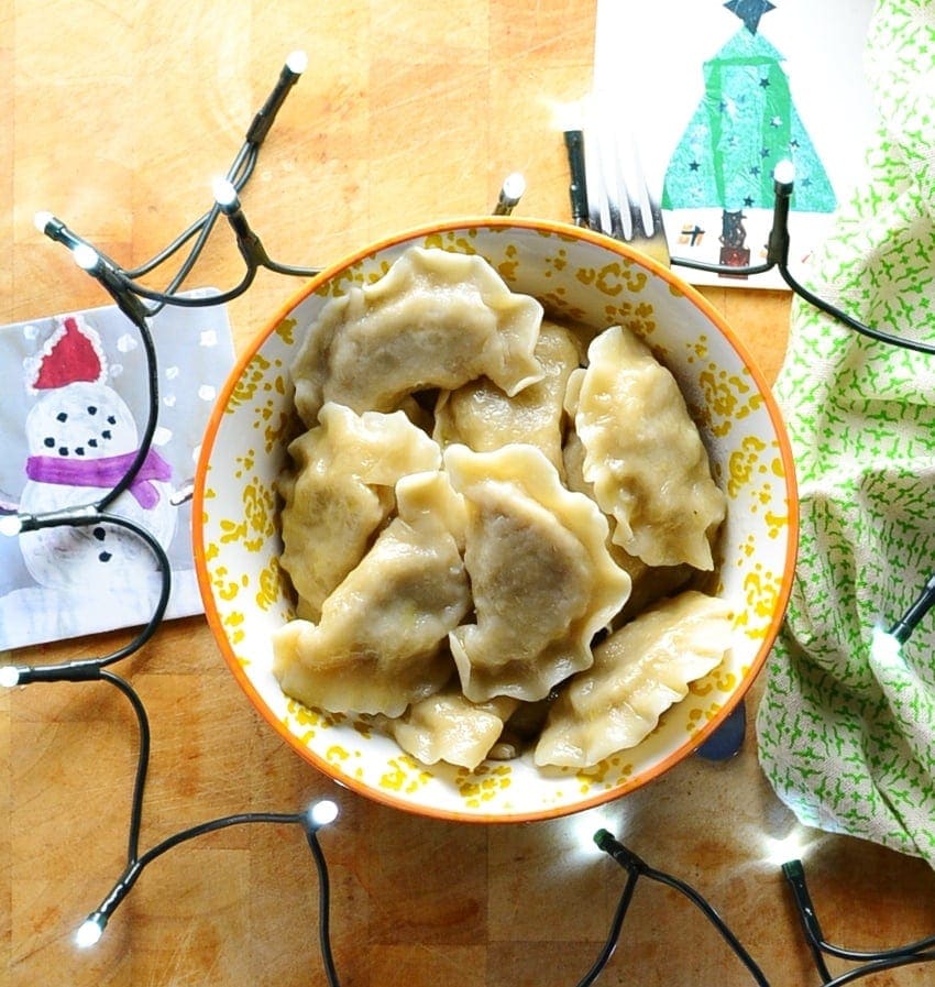 Sauerkraut pierogi in yellow-and-white patterned bowl with Christmas themed coasters, Christmas lights and green cloth on light wooden surface.