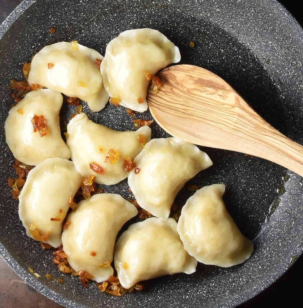 Top down view of cooked pierogi ruskie dumplings with crispy onion and wooden spoon in skillet.