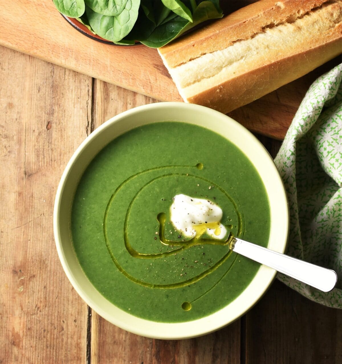 Creamy spinach soup with dollop of yogurt in green bowl with spoon, green cloth, baguette and spinach in background.
