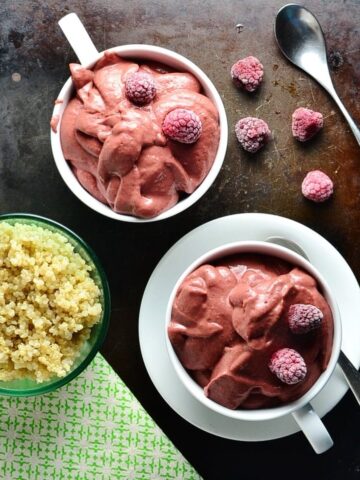 Top down view of raspberry smoothie in 2 white cups, one on saucer with spoon and frozen raspberries, with cooked quinoa in green dish and green cloth in bottom left corner.