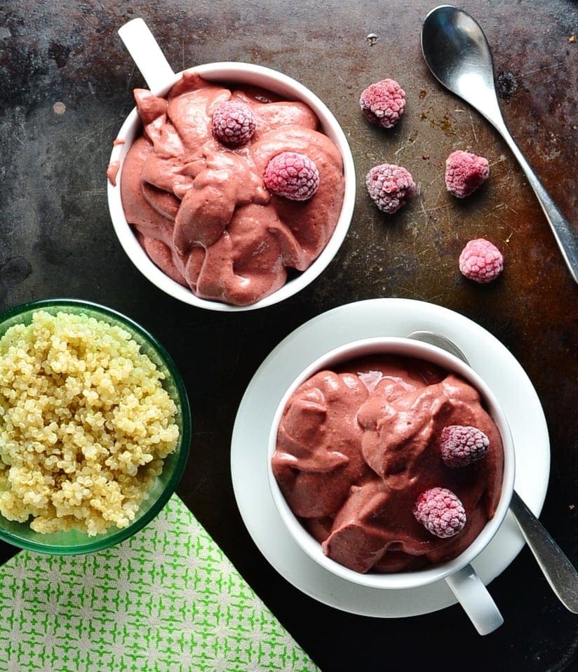 Raspberry smoothie in 2 white cups, one on saucer with spoon and frozen raspberries, with cooked quinoa in green dish and green cloth in bottom left corner.