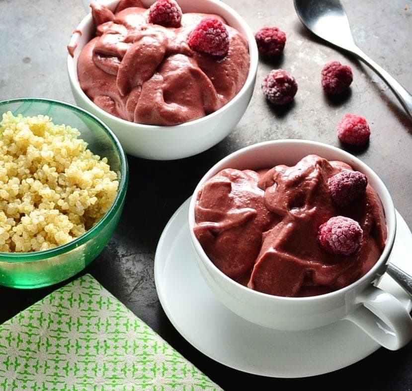 Pink quinoa smoothie in 2 white cups, one on saucer with spoon and frozen raspberries, with cooked quinoa in green dish and green cloth in bottom left corner.