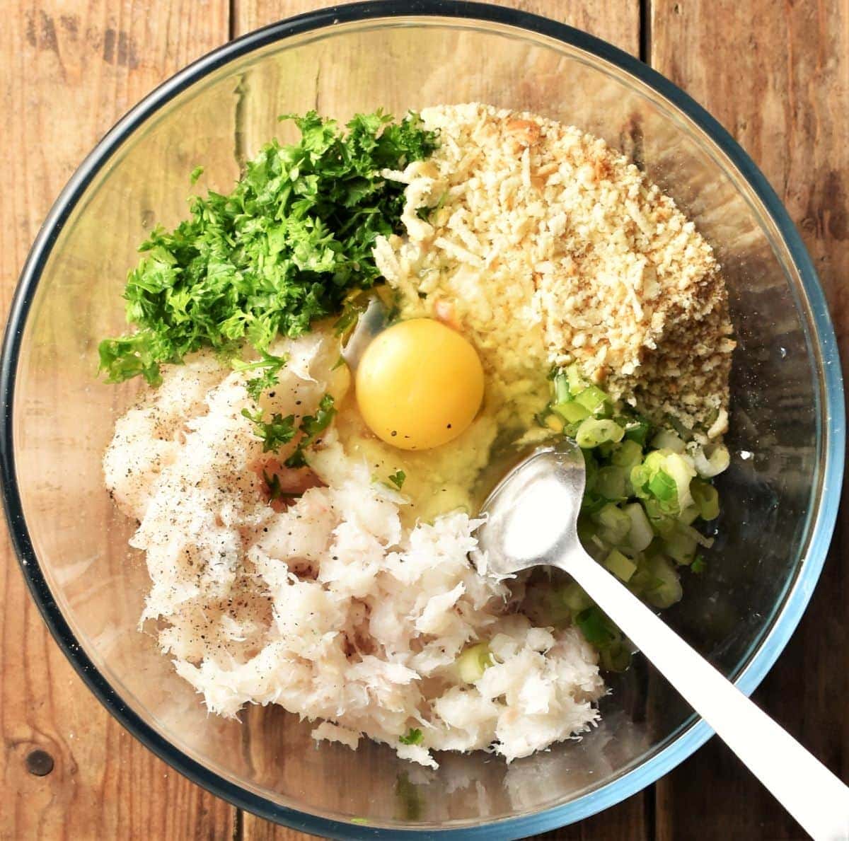 Fish, herbs, breadcrumbs and raw egg in mixing bowl with spoon.