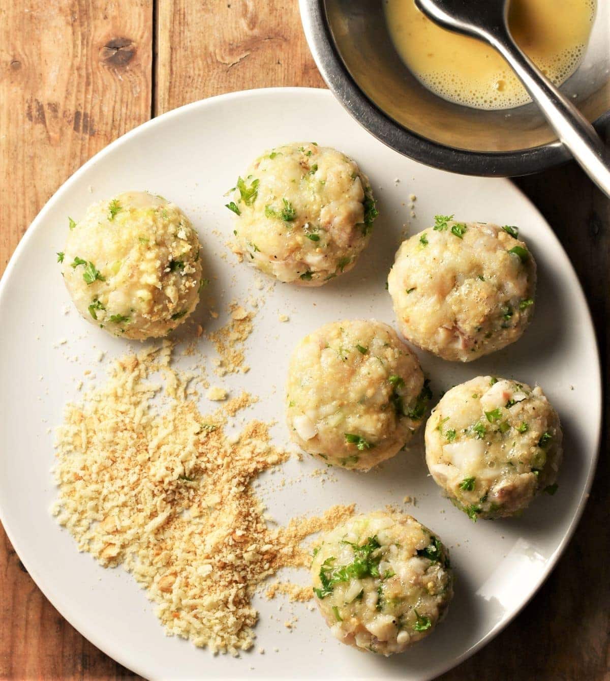 6 breaded fishcakes and breadcrumbs on top of plate with egg wash in top right.