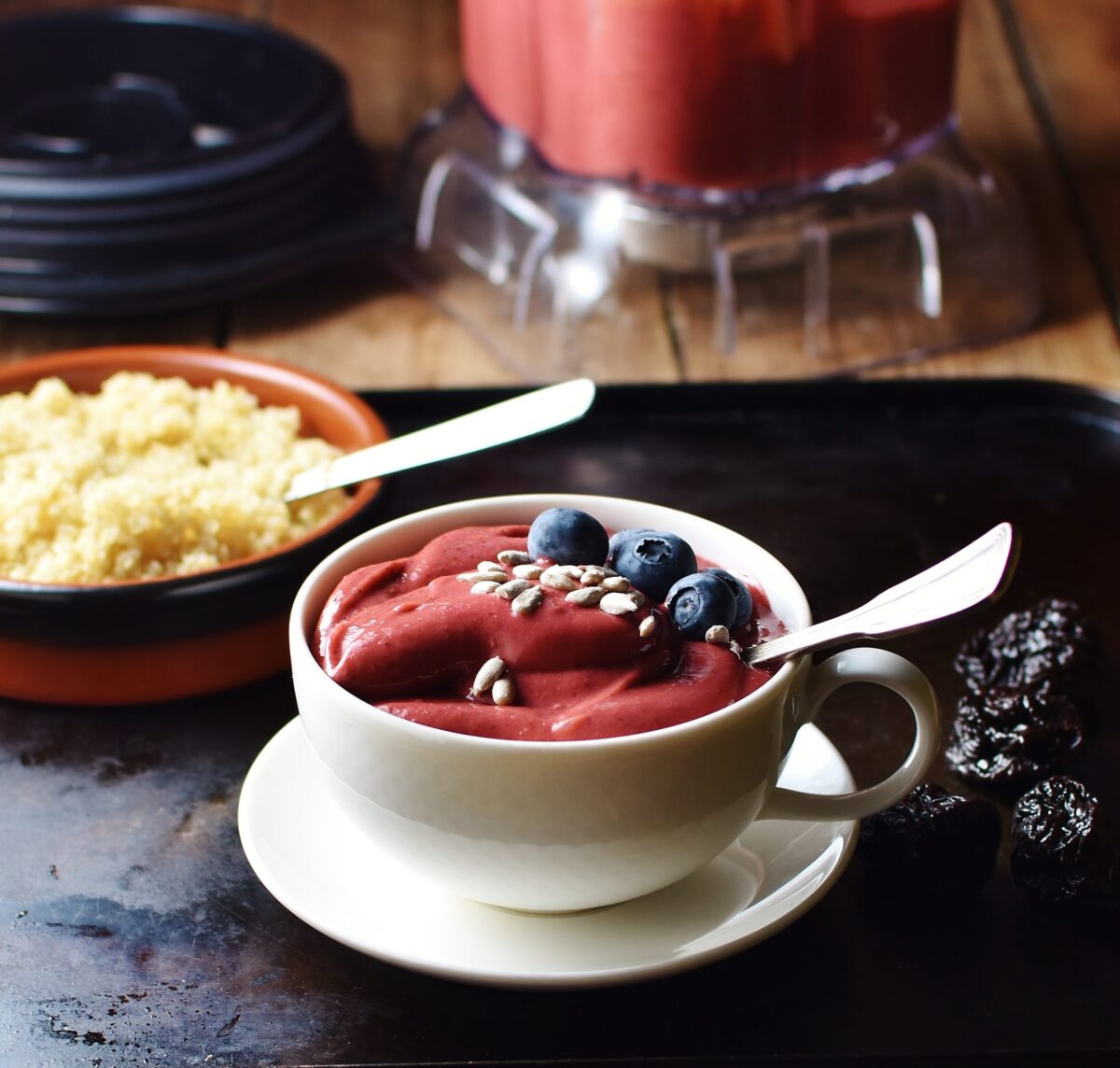 Side view of raspberry smoothie in white cup with spoon, seeds and blueberries on top of white saucer, with cooked quinoa in brown dish with spoon and smoothie in blender in background.