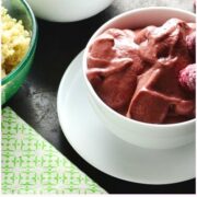 Raspberry smoothie in 2 white bowls with 1 saucer, fresh raspberries in background and green cloth in bottom left corner.
