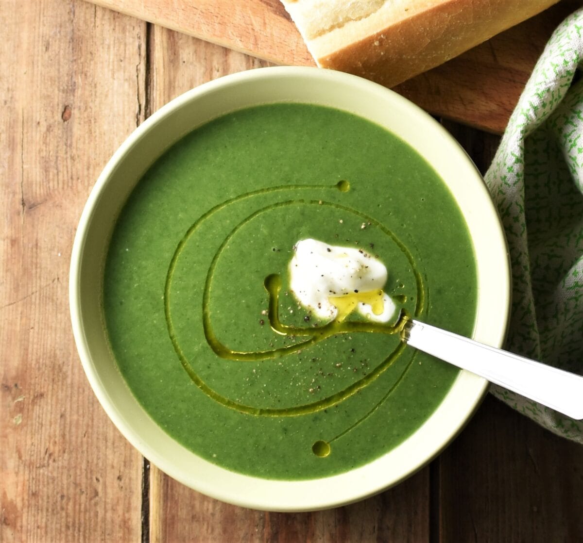 Cream of spinach soup in green bowl with spoon, baguette and green cloth in background.