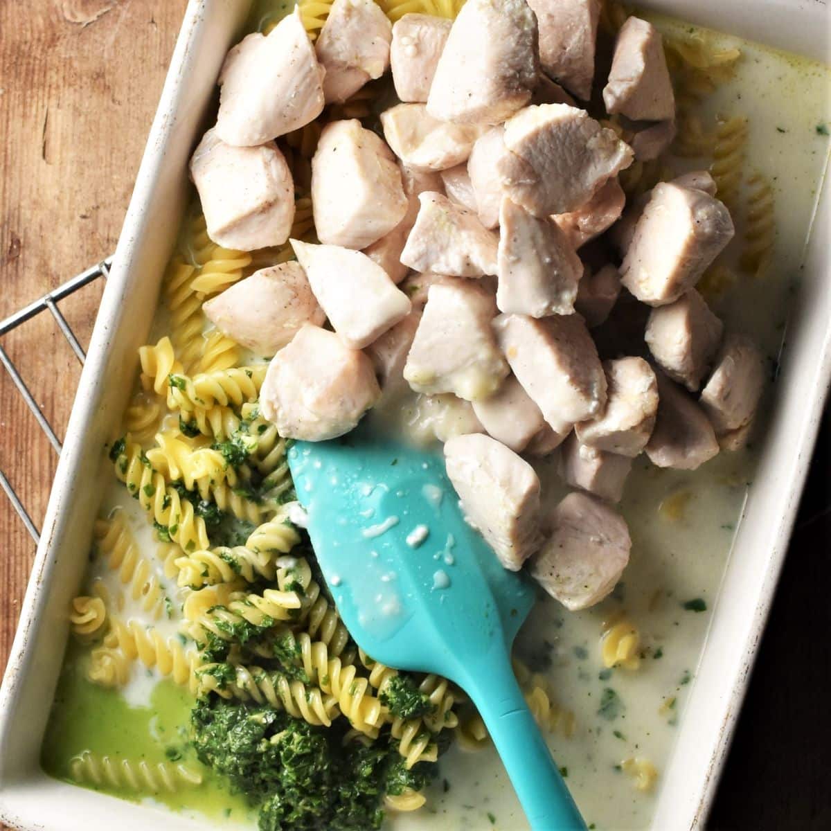 Assembling pesto pasta with chicken and creamy sauce in rectangular dish with blue spatula.