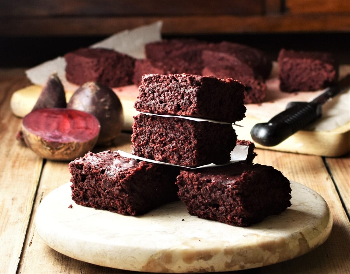 Side view of beetroot brownies on top of round marble plate, with raw beets and brownies in background.