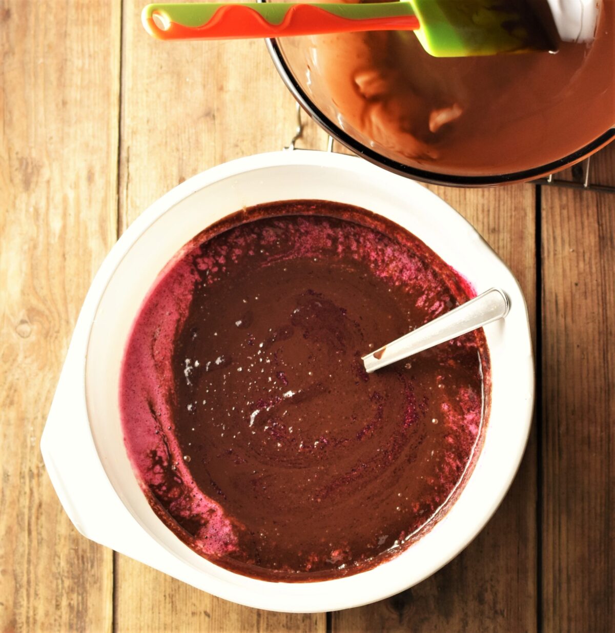 Chocolate beetroot brownie batter in a large white bowl with spoon and melted chocolate in bowl and green spatula in top right corner.