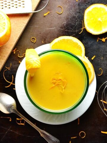 Orange sauce in bowl on white plate with cutting board, oranges, zester, spoon, whisk and orange zest on dark brown surface.