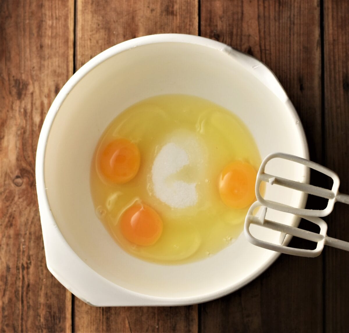 3 eggs and sugar in large white bowl with electric mixer end pieces.