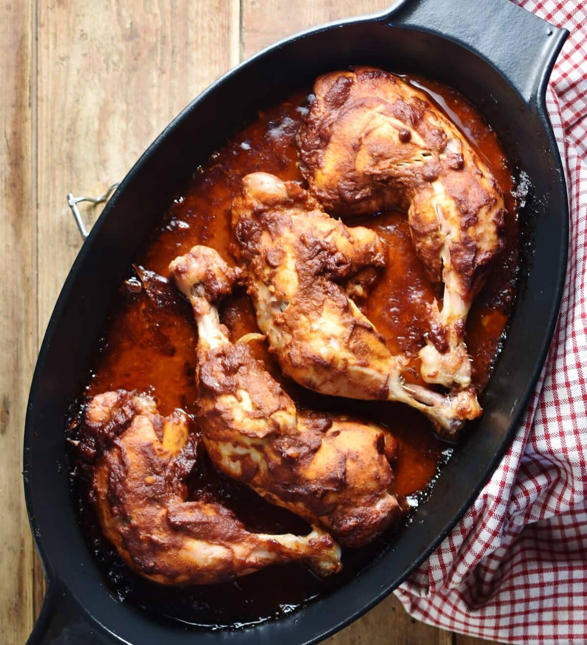 Cooked chicken legs in marinade inside oval black dish with red-and-white checkered cloth.