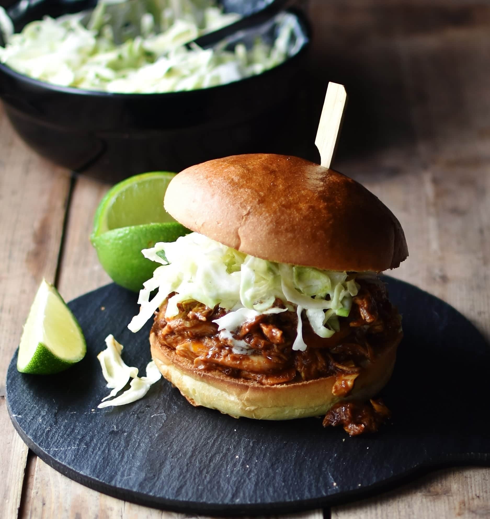 Side view of shredded chicken with coleslaw inside burger bun pierced with bamboos stick, with lime and coleslaw in black dish in background.