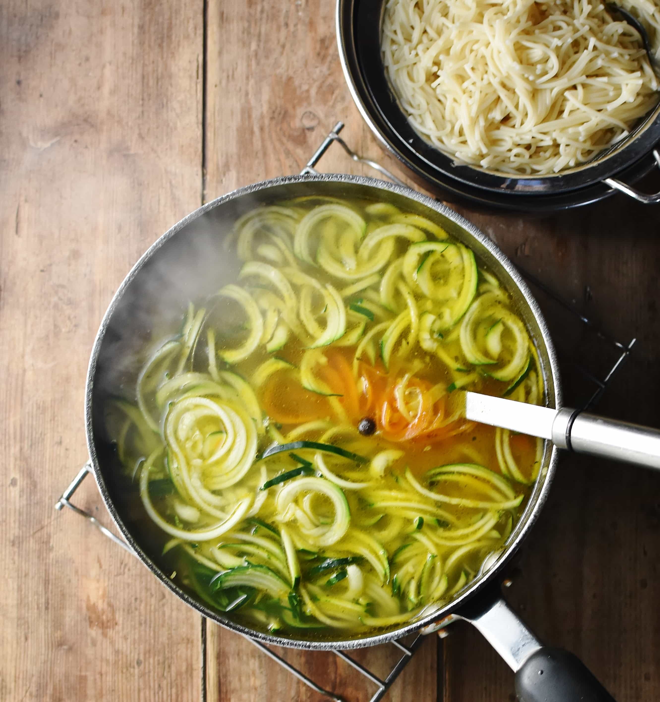 Zucchini noodles in hot soup with steam in large pot with ladle and cooked vermicelli pasta in top right corner.