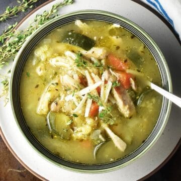 Top down view of chunky chicken zucchini soup in green bowl with spoon and thyme in background.