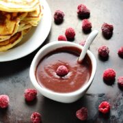 Chocolate sauce with raspberries in white bowl with spoon, with partial view of white plate with pancakes on dark surface.