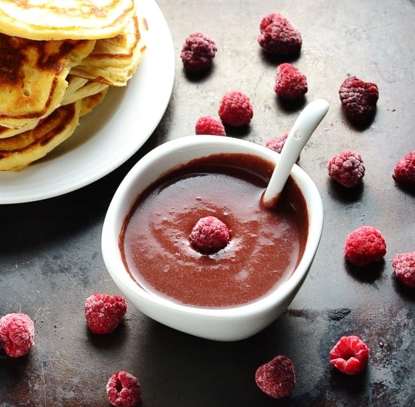 Chocolate sauce with raspberries in white bowl with spoon, with partial view of white plate with pancakes on dark surface.