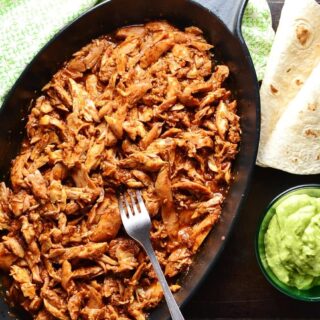Top down view of pulled chicken in black oval cast iron dish with fork, wraps, guacamole in small dish and green cloth.