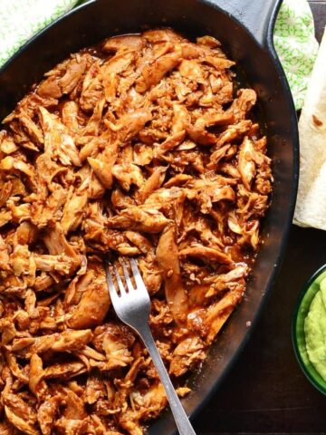 Top down view of pulled chicken in black oval cast iron dish with fork, wraps, guacamole in small dish and green cloth.