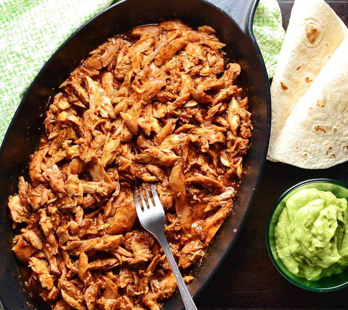 Top down view of pulled chicken in black oval cast iron dish with fork, wraps, guacamole in small dish and green cloth.