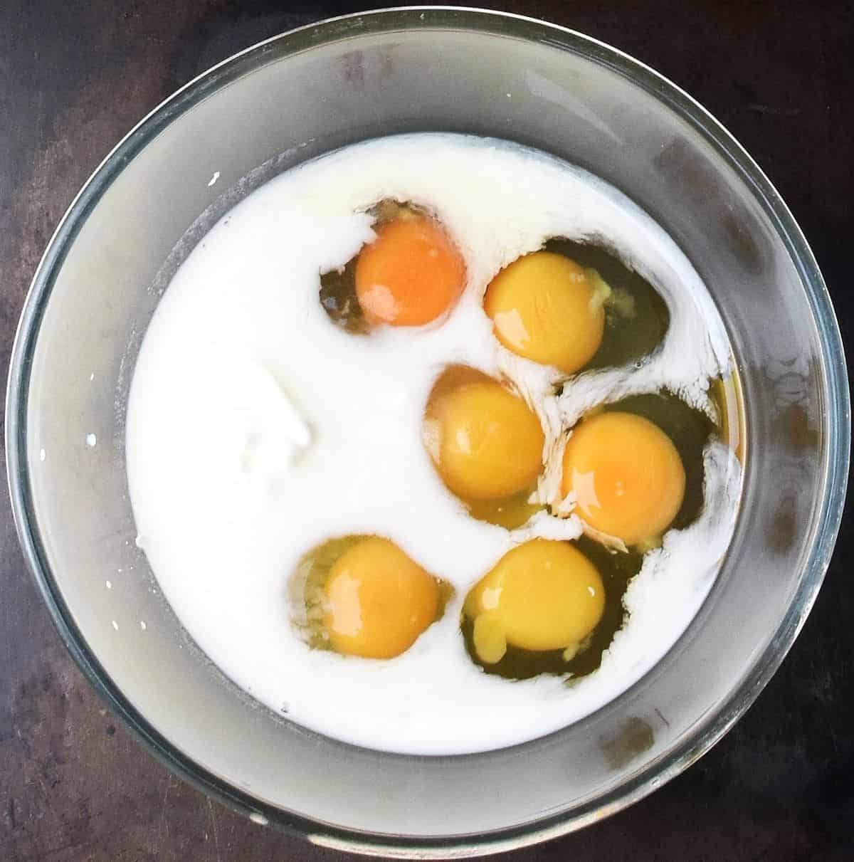 Six raw eggs with milk and yogurt in glass bowl.